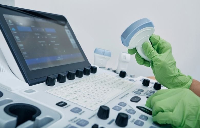 Eine Hand in grünem Handschuh hält einen Schallkopf vor dem Bedienfeld eines Ultraschallgeräts.