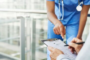 Eine medizinische Fachkraft zeigt auf einen Fragebogen zur Patientenaufnahme, den ein Patient auf einem Klemmbrett ausfüllt.