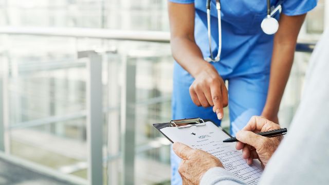 Eine medizinische Fachkraft zeigt auf einen Fragebogen zur Patientenaufnahme, den ein Patient auf einem Klemmbrett ausfüllt.