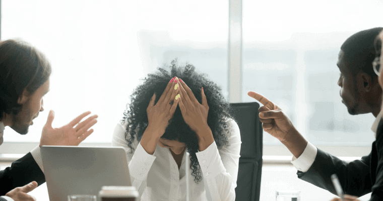 A person of color puts her head down at an office table while other leaders point the finger.