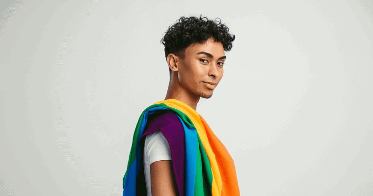 A LGBTQIA+ person looks knowingly at the camera with a rainbow flag draped over his shoulder.