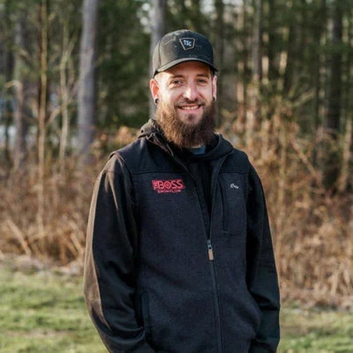 Man smiling outdoors in a forest setting, wearing a black jacket and cap.
