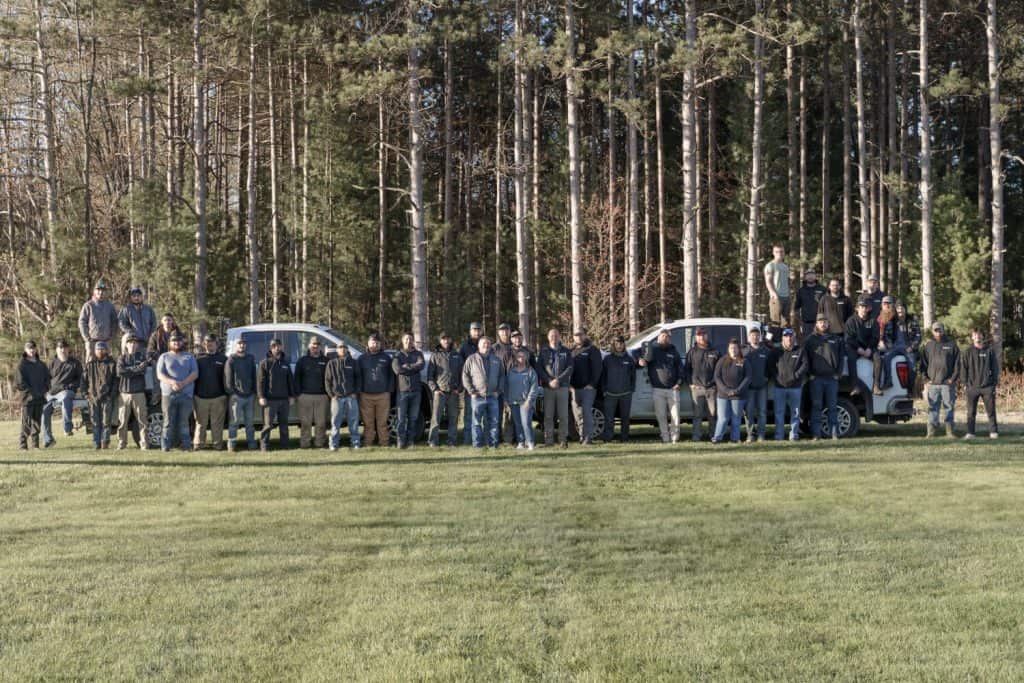 People standing in a line with cars parked behind them, surrounded by trees.