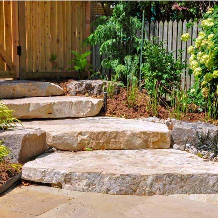 Stone garden steps with lush greenery and wooden fence in the background.