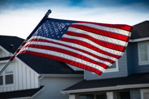 A United States flag is waving in front of a suburban house under a clear Bentonville sky, symbolizing the community spirit fostered by Bentonville Mayor Stephanie Orman.