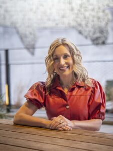 Bentonville Mayor, Stephanie Orman in a red shirt with puffy sleeves sits at a wooden table, smiling, with a blurred background of a local outdoor art piece and a wall in Bentonville.