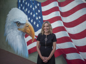 A woman in a black dress stands smiling in front of a mural featuring a bald eagle and a waving American flag.