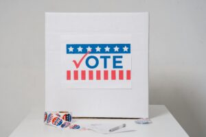 A voting booth with a "Vote" sign displaying stars and stripes. Nearby, a pen and a roll of "I Voted" stickers are on a white table.
