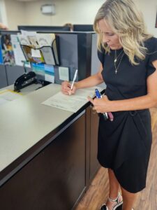 A woman standing at a counter fills out a form. She is holding multiple pens and wears a black dress and white sandals. Office supplies and dividers are visible in the background.