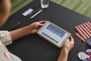 A person holds a tablet showing an electronic voter registration form. On the desk are an American flag, a voter ID card, a pen, and voter buttons.