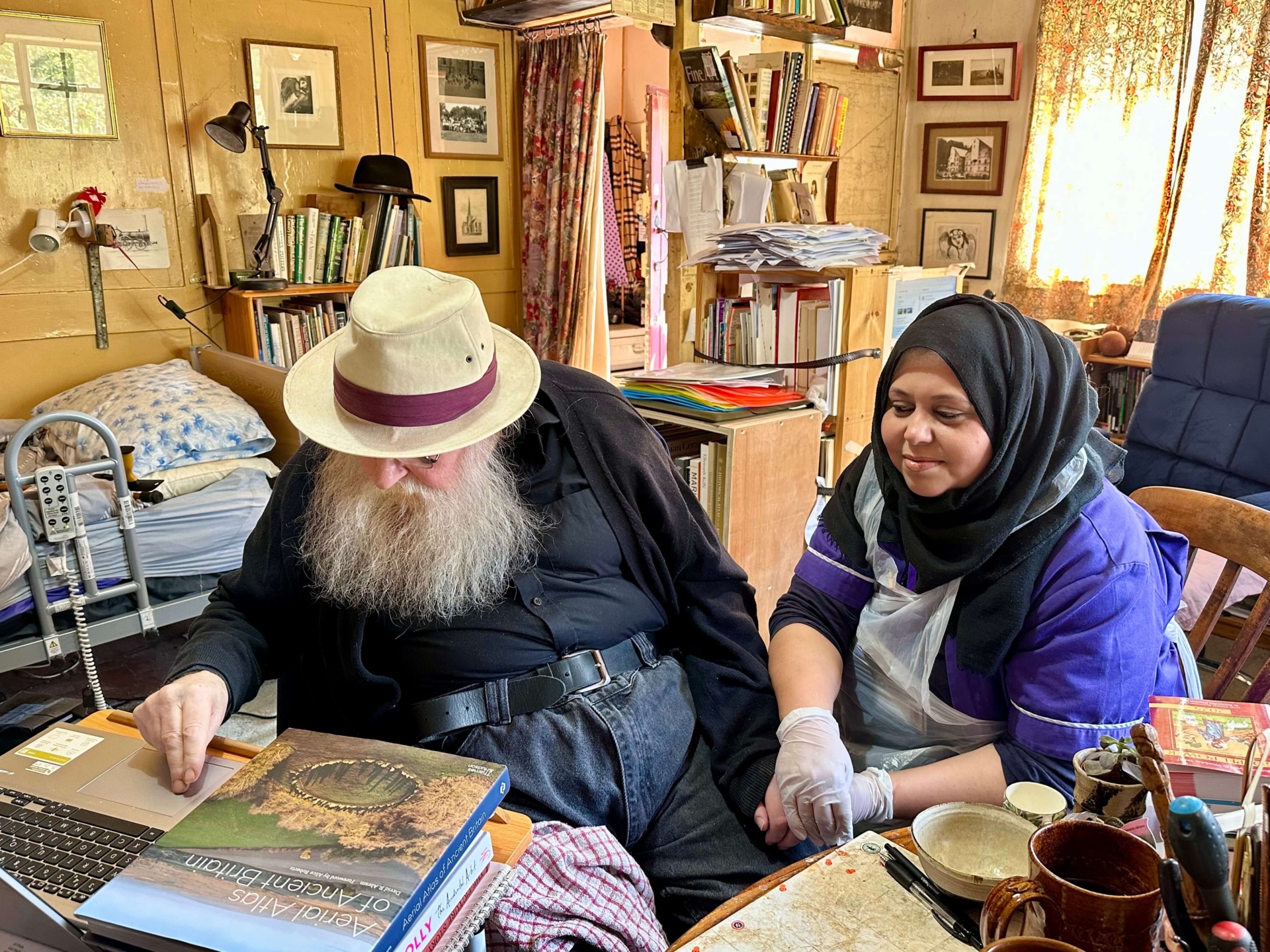 An elderly man with a white beard and hat uses a laptop at a cluttered desk, while his live-in care companion, a woman in a hijab and gloves, sits beside him holding his hand in their cozy, book-filled room with warm lighting.