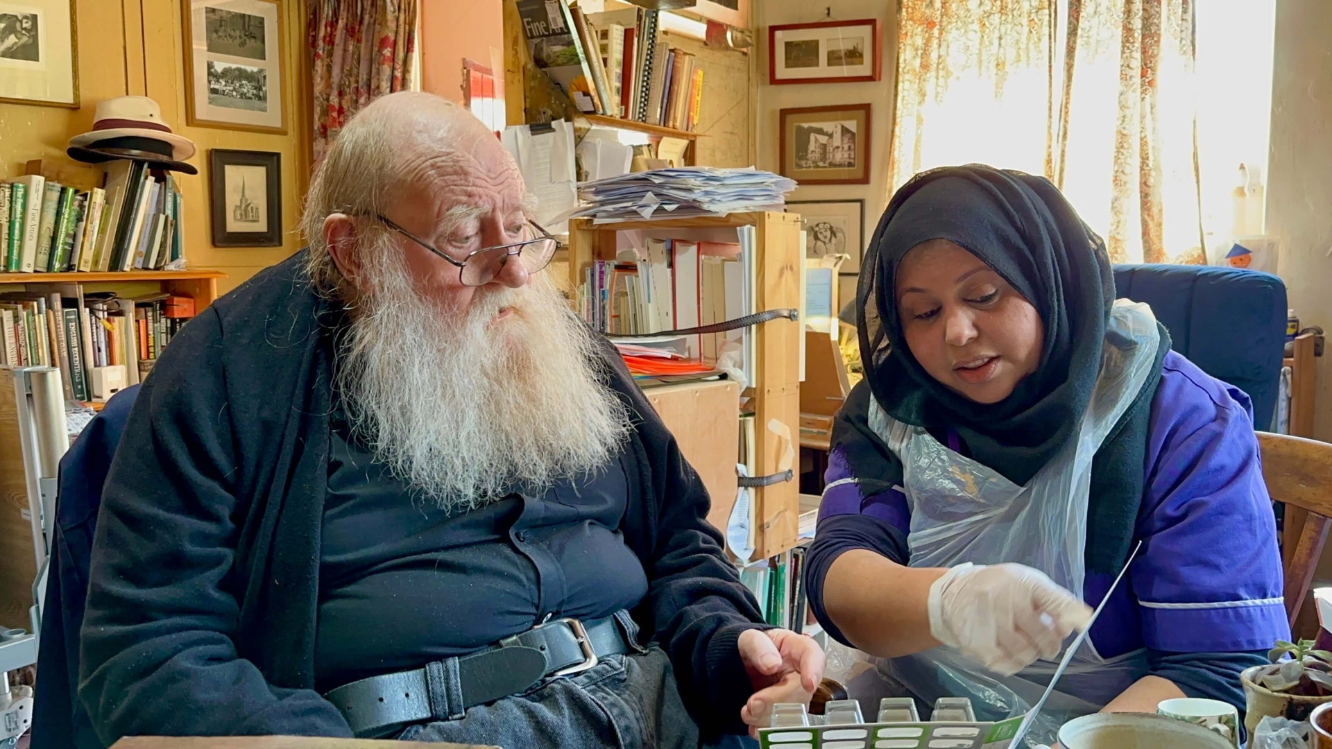 An elderly man with a long white beard sits beside a younger woman wearing a hijab and gloves. With the support of live-in care, they enjoy gardening together, planting seeds in pots on a table in a cozy, book-filled room.