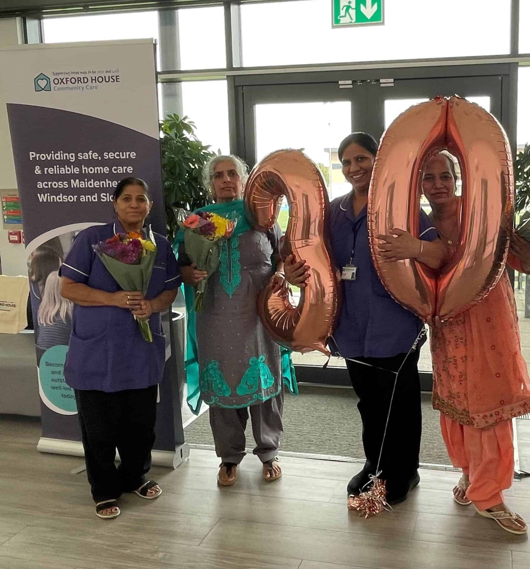 Four individuals smiling and celebrating, with two holding flower bouquets and two holding balloons shaped as the numbers "9" and "0," possibly marking a special occasion or anniversary.