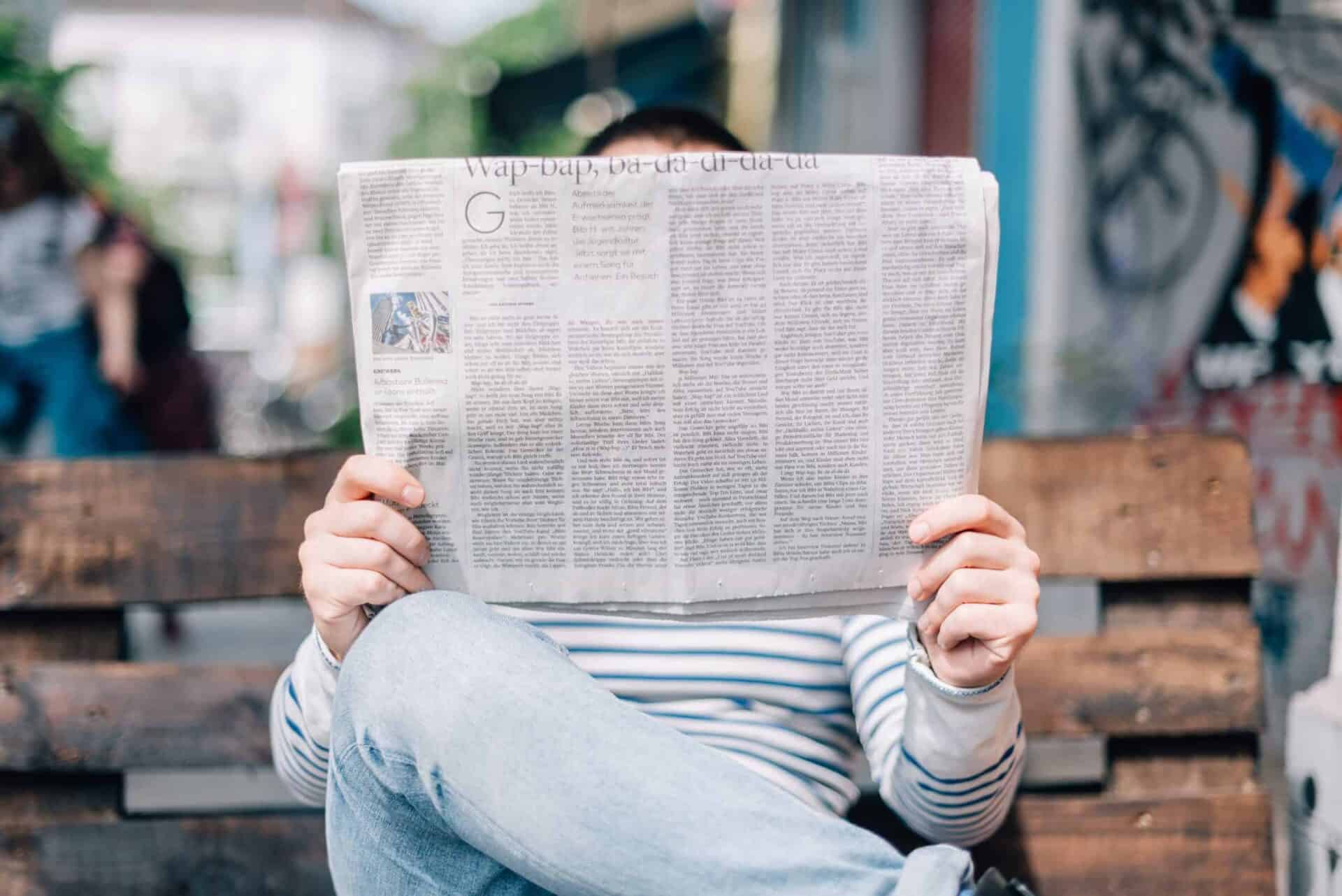 A person sitting on a wooden bench outside while reading a newspaper.