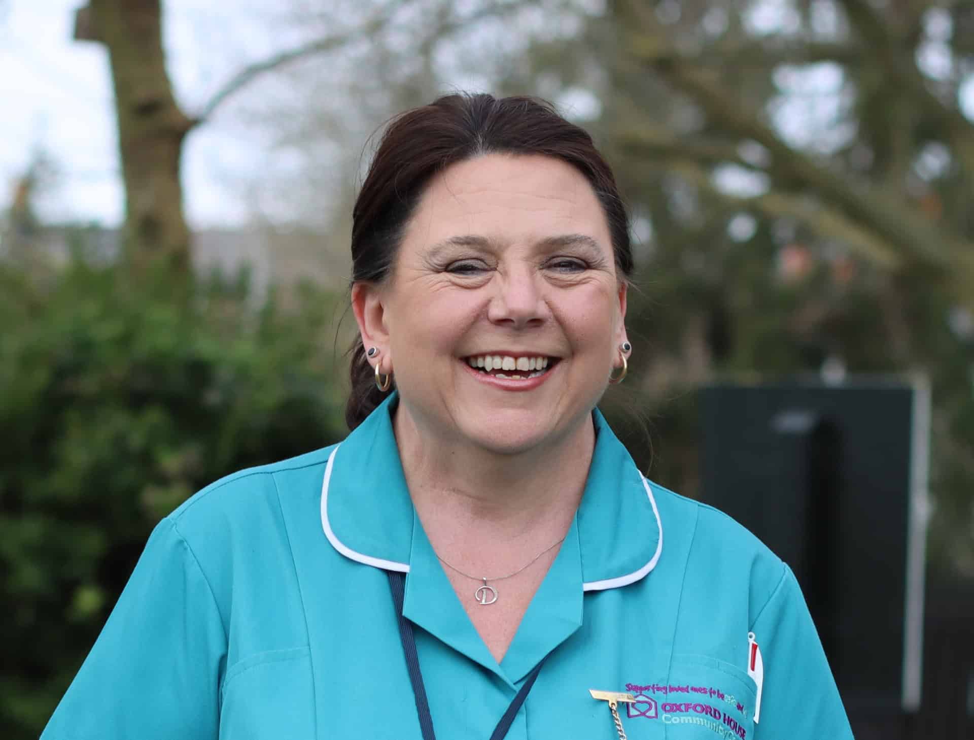 A woman wearing a teal nurse uniform smiles outdoors. Trees and greenery are faintly visible in the background.