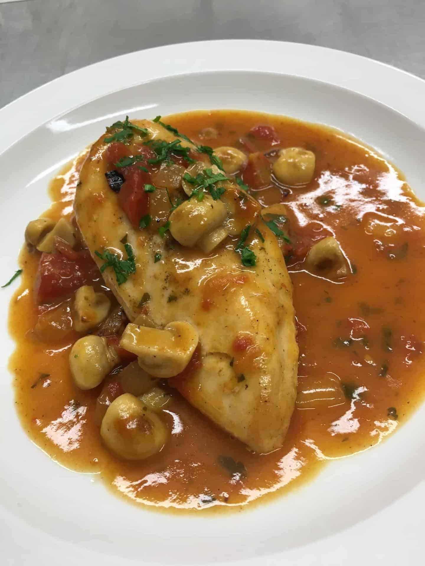 A plate of chicken breast topped with a tomato-based sauce, garnished with sliced mushrooms and herbs. The dish is served on a white plate, showcasing a rich, colorful presentation.