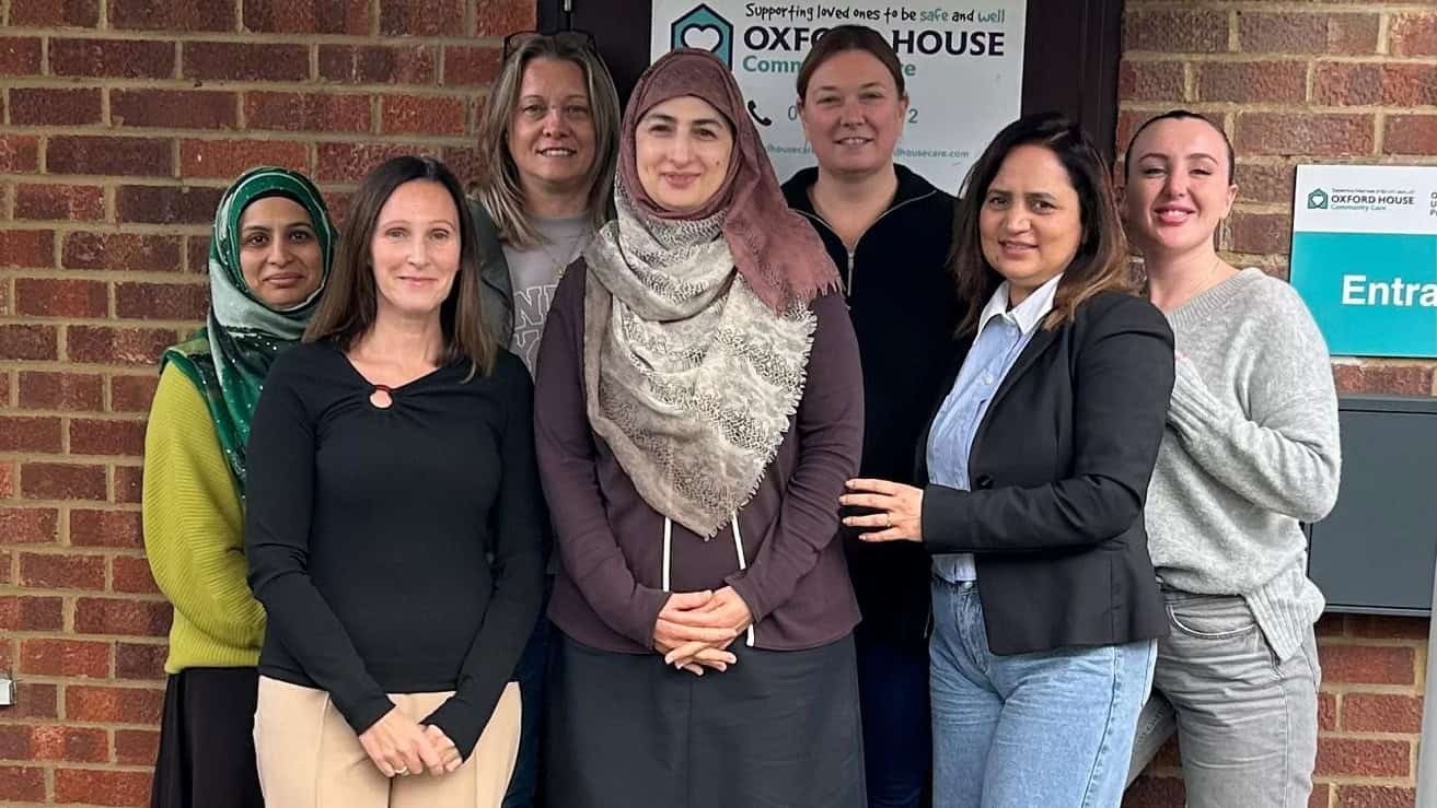 A group of seven people stands in front of a brick building with a sign that reads "Oxford House." They are smiling and casually dressed, with some wearing scarves and jackets.