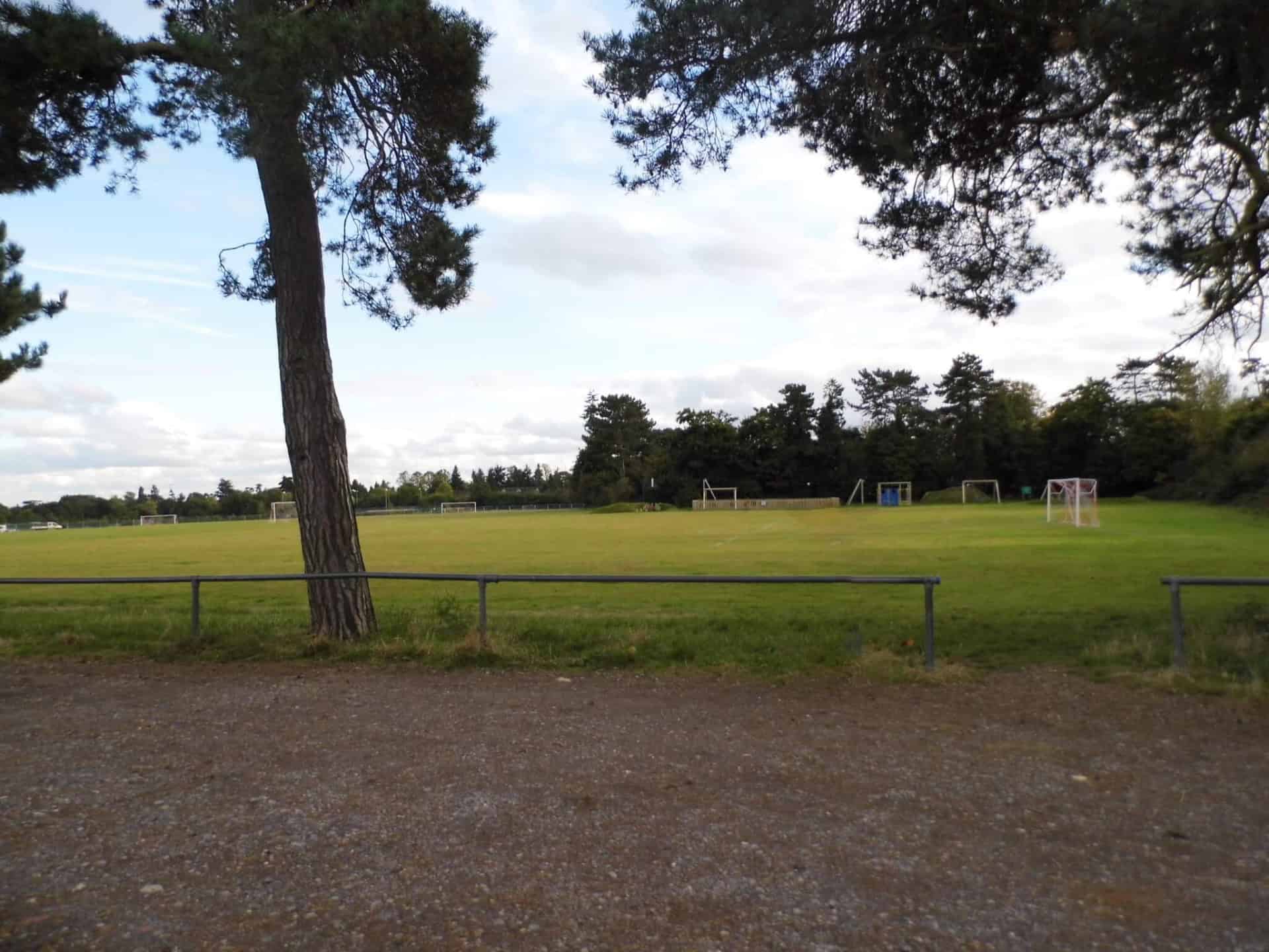 A grassy sports field with several football goals is surrounded by trees in Farnham Royal. A dirt path with a green trash bin runs along the foreground. The sky is partly cloudy, and tall, scattered trees frame parts of the scene.