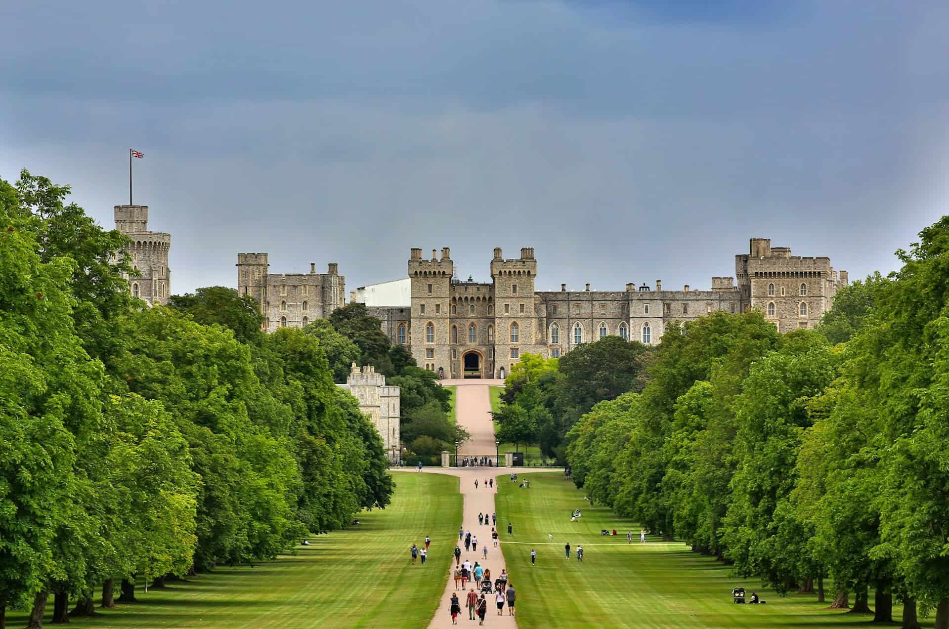 A grand castle with multiple towers is surrounded by lush greenery. A wide, tree-lined path leads up to the castle's entrance. People are walking along the pathway under a cloudy sky.