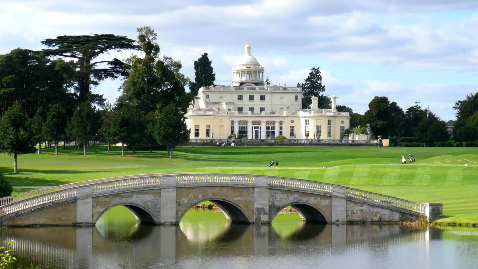 Elegant mansion in Stoke Park with a white facade and dome, set in a lush landscape with green lawns and large trees. A stone arch bridge spans a reflective body of water in the foreground, while the sky is partly cloudy.
