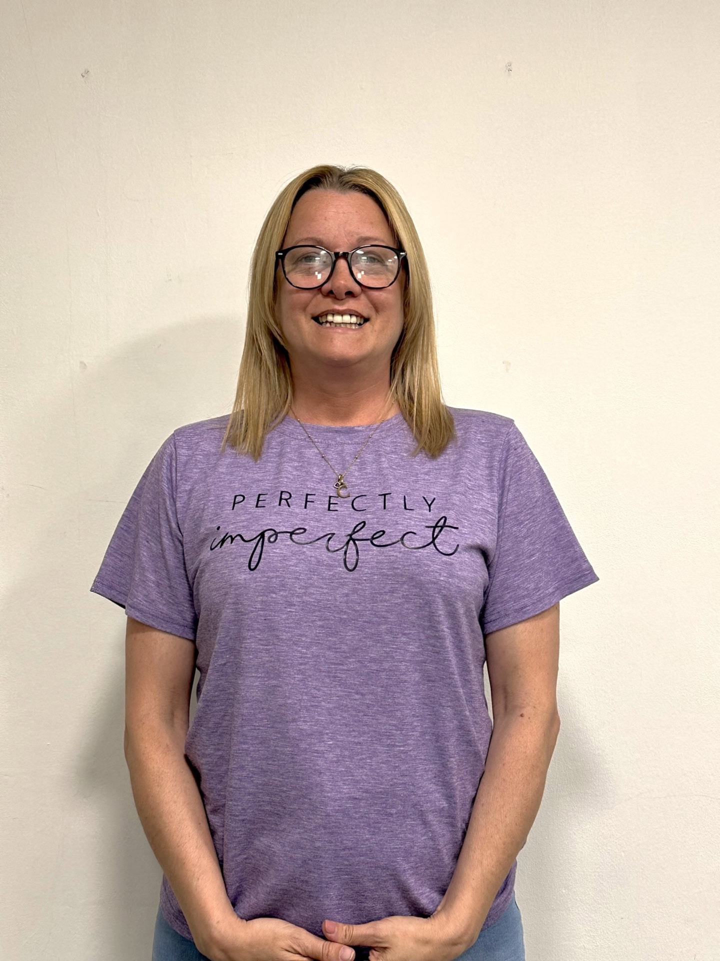 Meet our team: A woman with straight blonde hair and glasses smiles at the camera, wearing a purple T-shirt that reads "Perfectly imperfect," standing against a plain, light-colored wall.