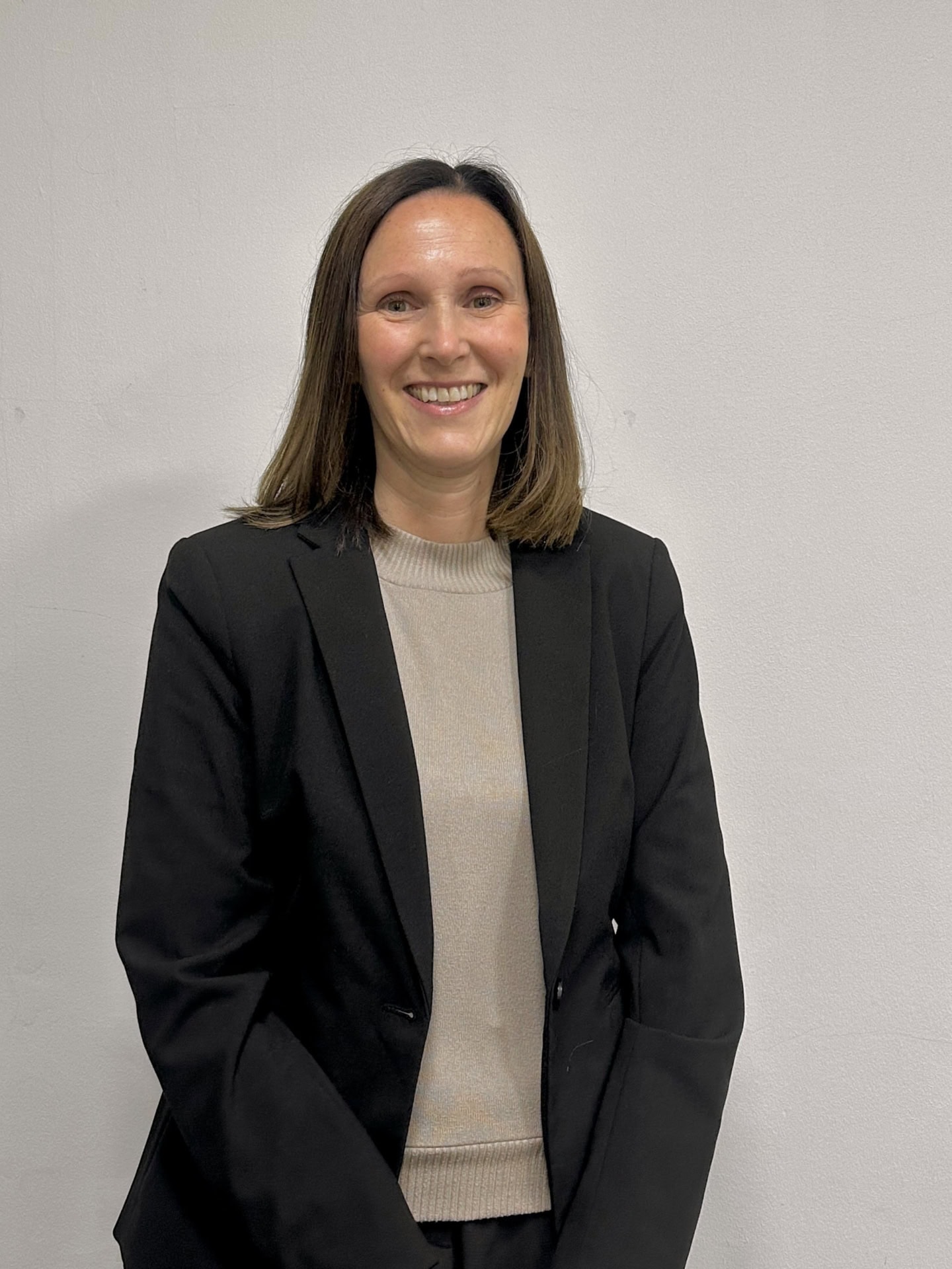 Meet our team: A woman with straight brown hair, wearing a black blazer over a light beige top, smiles while standing in front of a plain white wall.