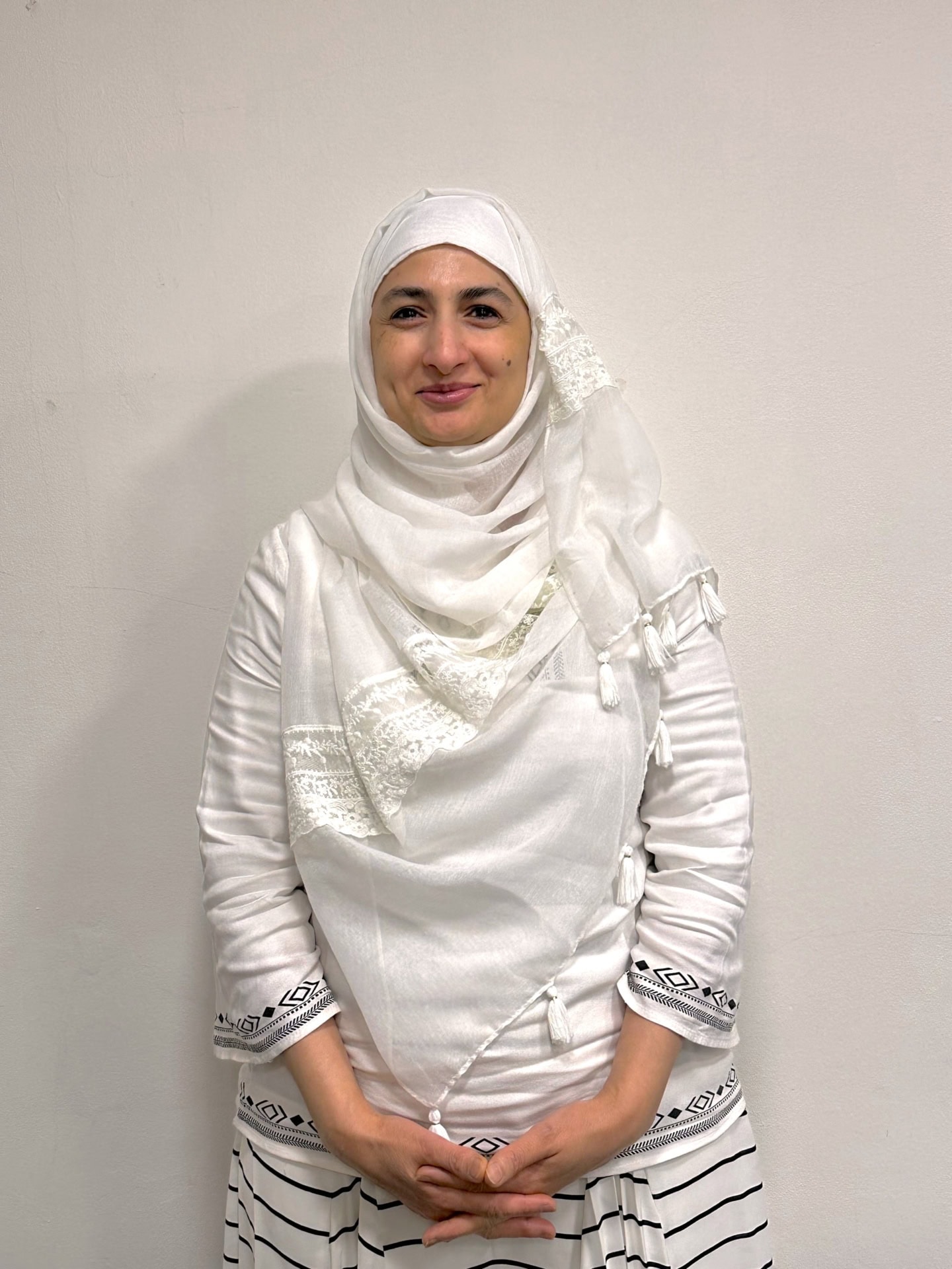 Meet our team: A woman wearing a white hijab and a white dress with black patterns on the sleeves and skirt stands against a plain light-colored wall, smiling gently with her hands clasped in front of her.