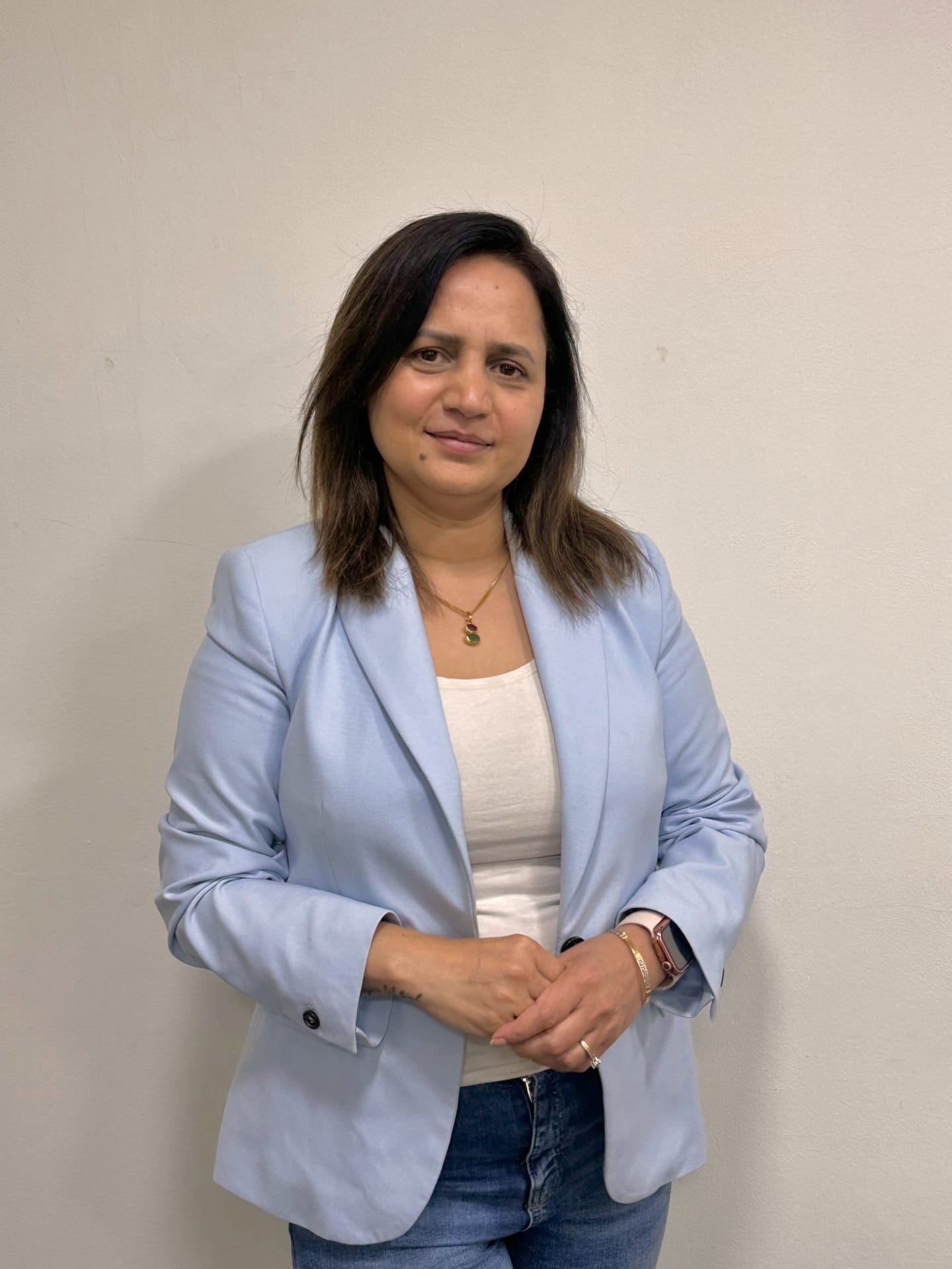 Meet our team: A woman with medium-length dark hair stands against a plain light-colored wall, wearing a light blue blazer, white top, and jeans. She is smiling slightly and has her hands loosely clasped in front of her.