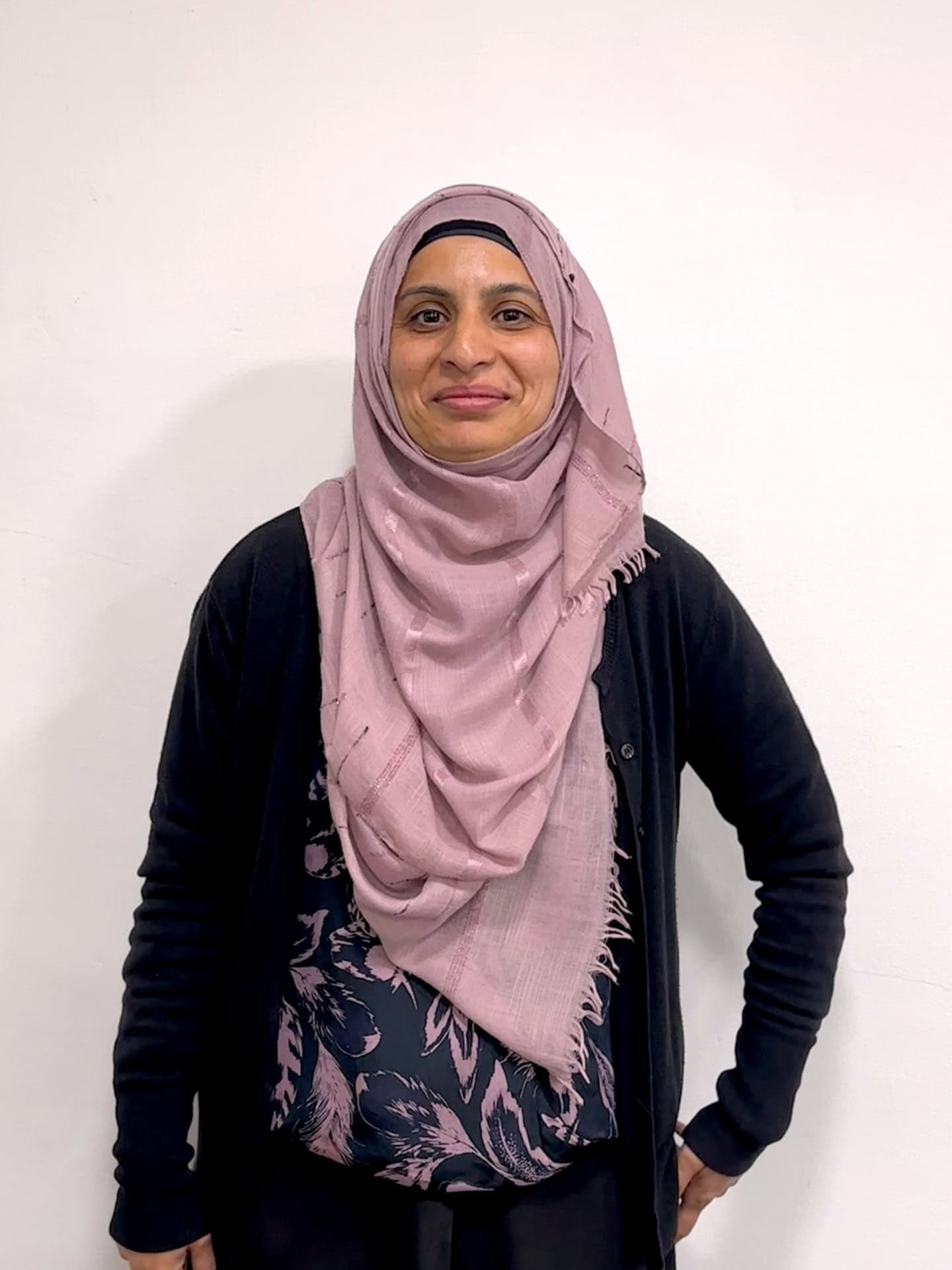 Meet our team: A woman wearing a light pink hijab, a dark cardigan, and a floral-patterned top stands smiling in front of a plain white wall.