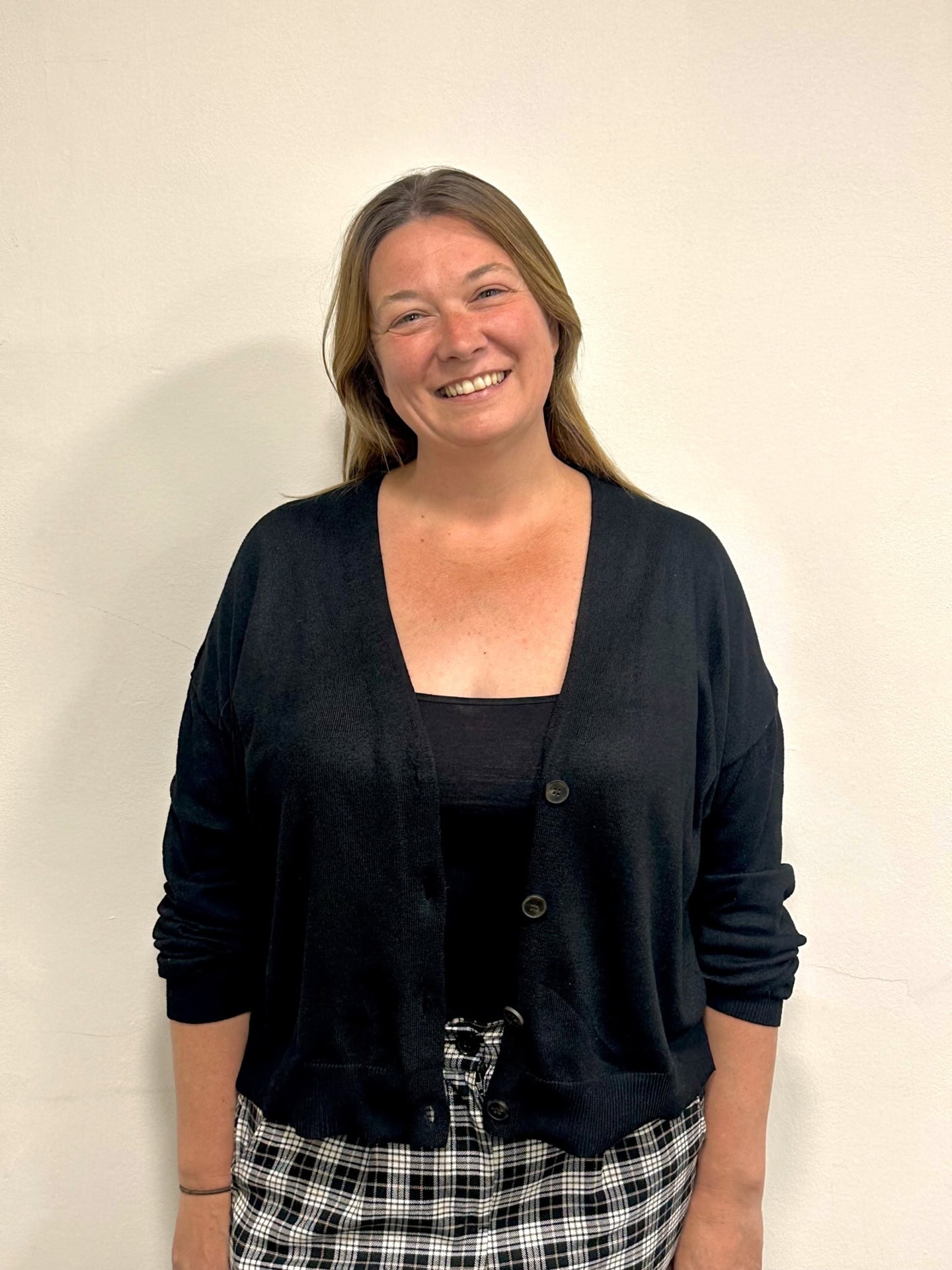 Meet our team: A woman with light brown hair smiles at the camera, wearing a black cardigan over a black top and plaid pants as she stands in front of a plain, light-colored wall.