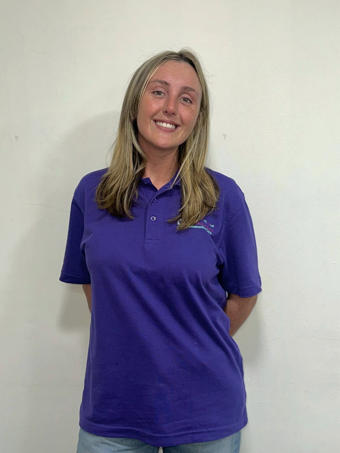 Meet our team: A woman with long, light brown hair is smiling and standing against a plain white wall. She is wearing a purple polo shirt and has her hands behind her back.