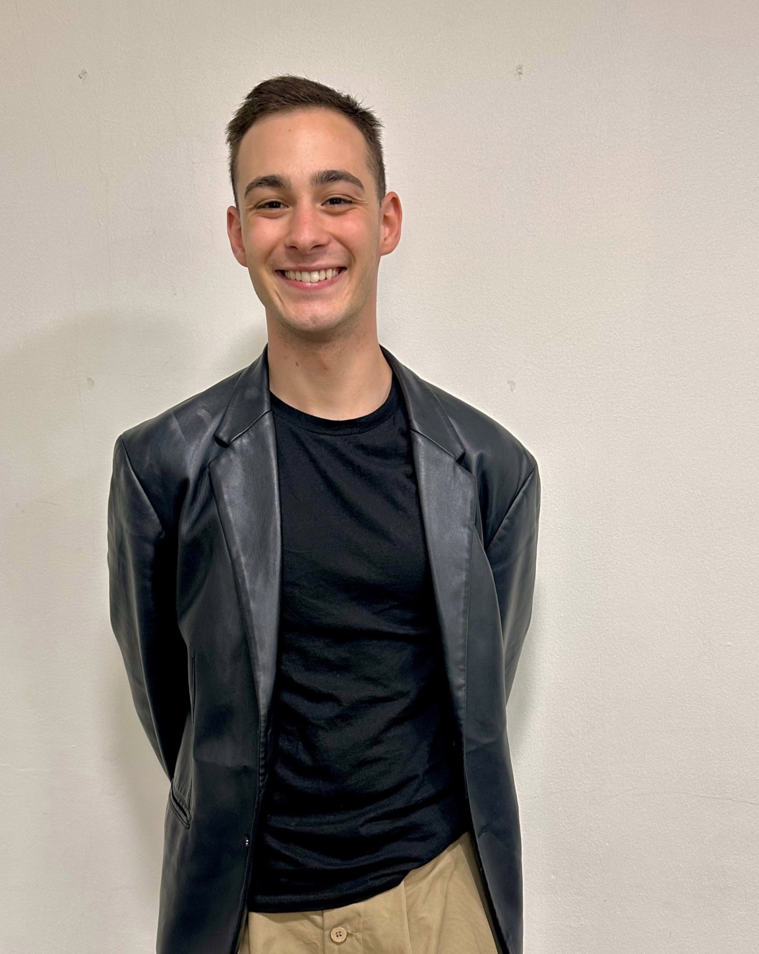 Meet our team: A young man smiling, wearing a black t-shirt, black leather jacket, and khaki pants, standing against a plain off-white wall.