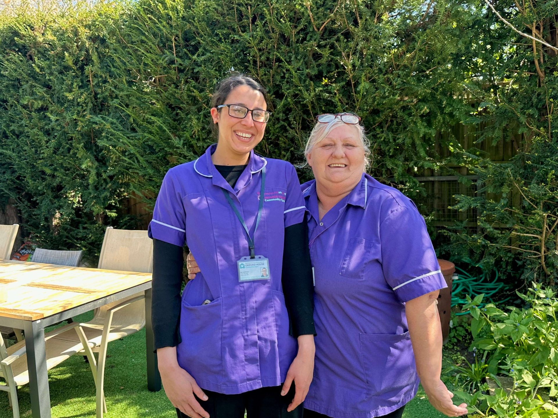 Two women in purple uniforms stand smiling together outside in a sunny garden, celebrating Good Care Month, with a wooden table and chairs nearby and green bushes in the background.