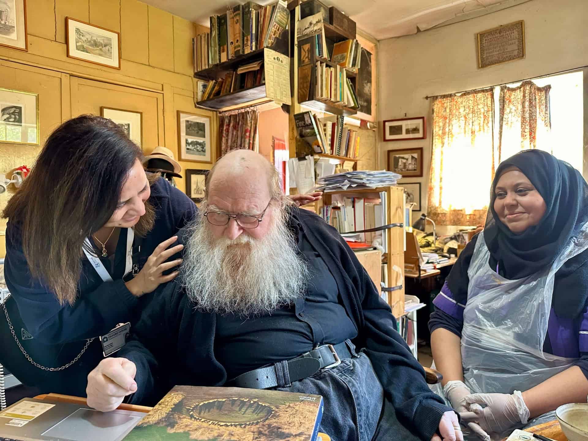 Three people in a cluttered, book-filled room. An elderly man with a long white beard sits between two women, one smiling and gently touching his shoulder, the other seated with hands in gloves and wearing a headscarf and apron.