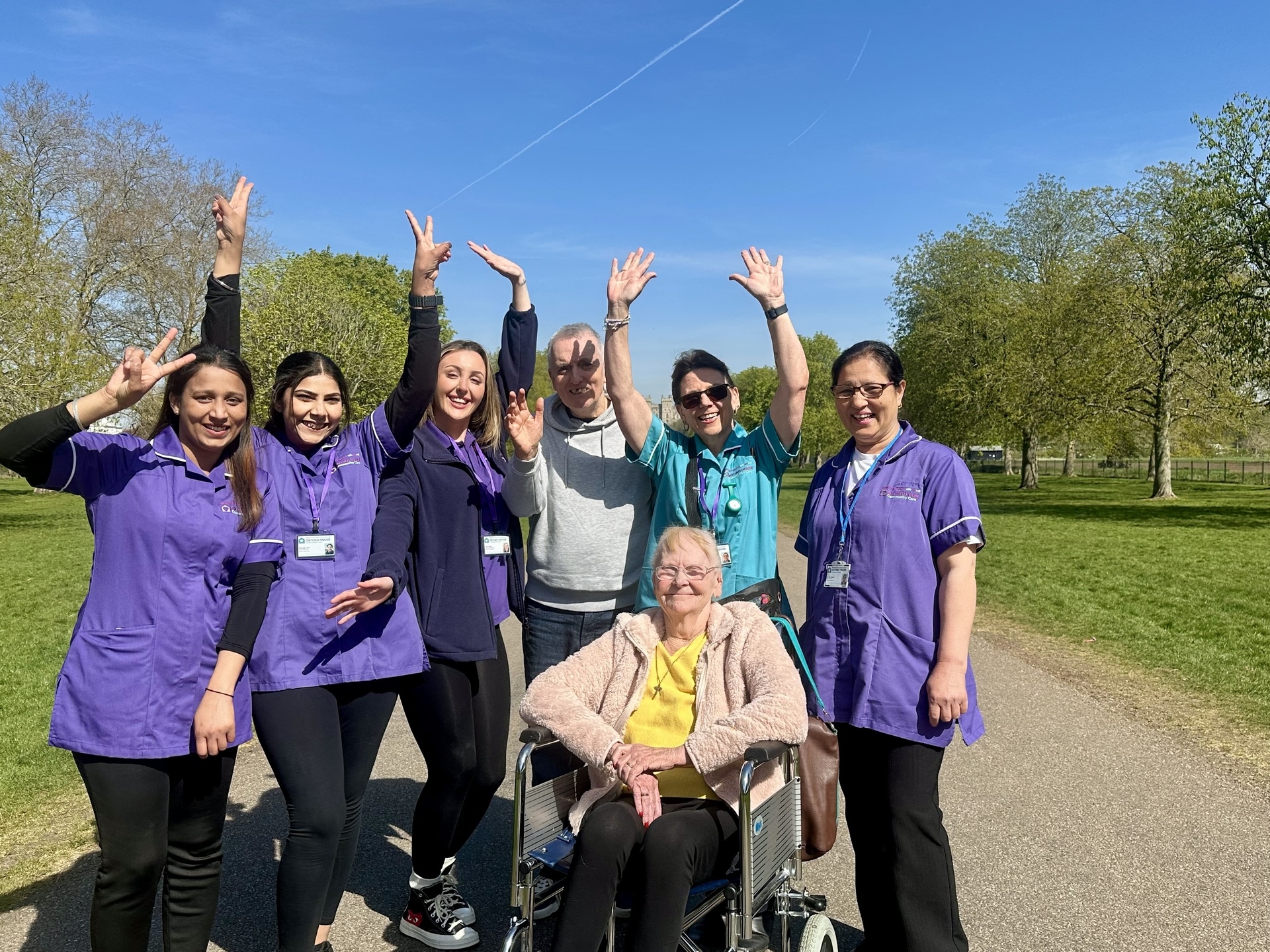 A group of smiling people, including four Oxford House caregivers in purple uniforms, two adults, and an elderly woman in a wheelchair, stand on a sunny path with trees and blue sky. Everyone looks happy and cheerful, reflecting quality home care.