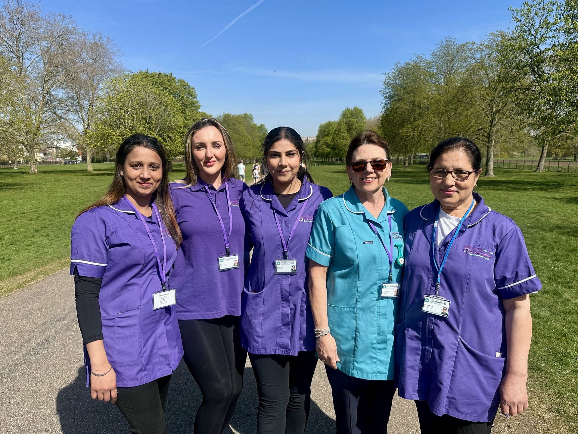 Five women, four in purple uniforms and one in teal, stand together smiling in a park on a sunny day. They wear name badges and lanyards, with trees and grass visible in the background.