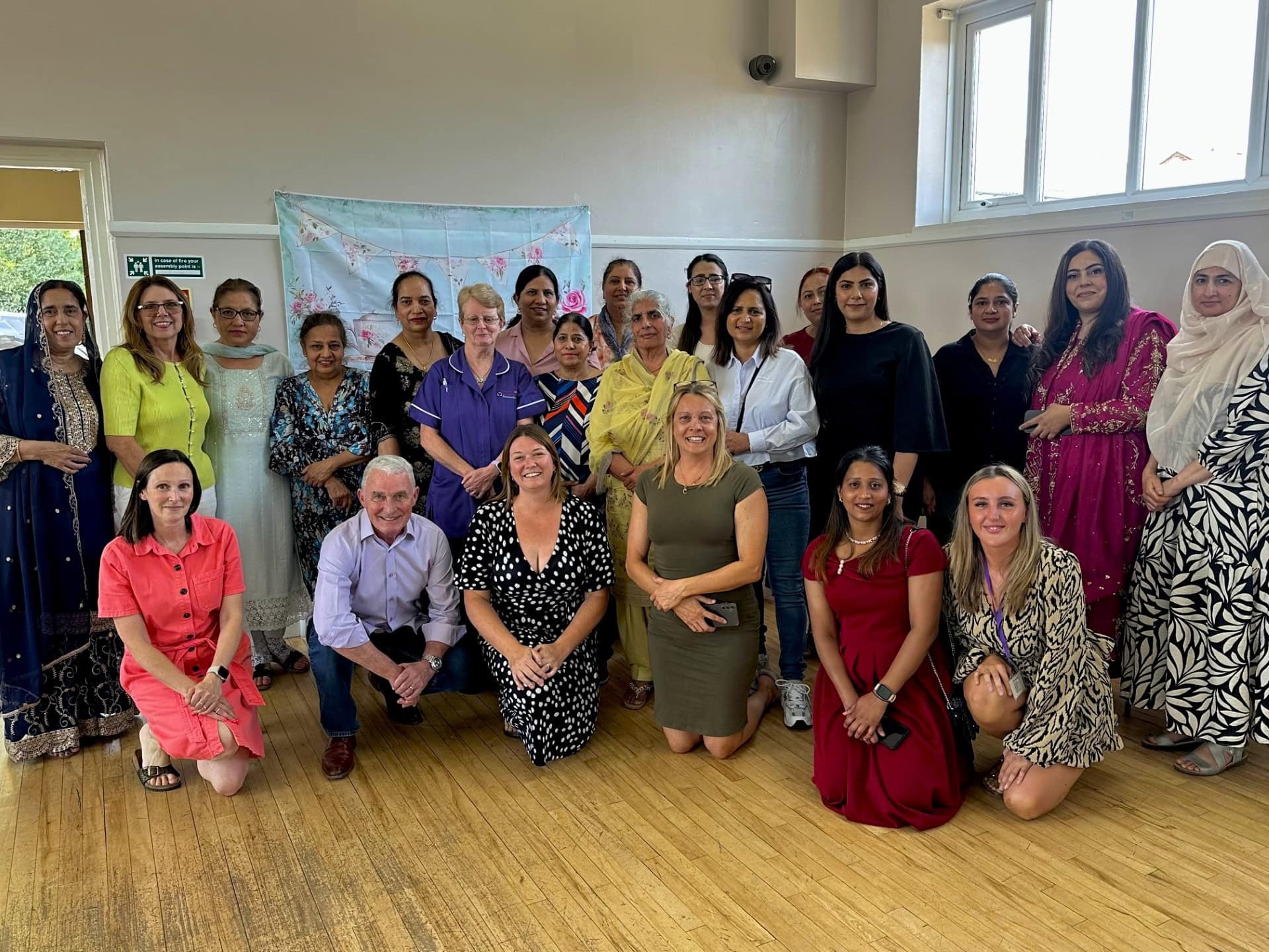 A diverse group of men and women pose and smile together indoors; most are standing, a few are kneeling in front. They are dressed in colorful and varied outfits, in a well-lit room with wooden floors.