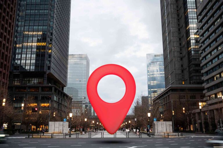 A large red map marker icon is placed in the middle of an urban street lined with high-rise buildings at dusk.