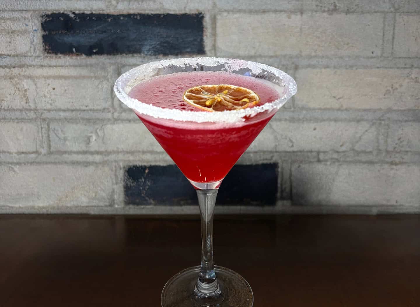 Bright red cocktail in a sugar-rimmed martini glass with a dried citrus garnish.