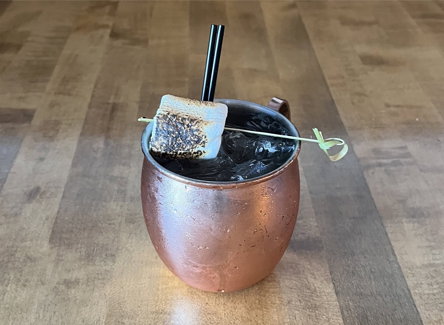 Copper mug with dark cocktail, marshmallow, and straw on wooden table.