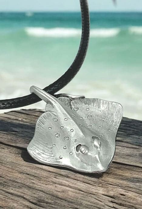 Un pendentif en étain massif représentant une raie pastenague évoque l'élégance marine sur fond de plage exotique. #Océan