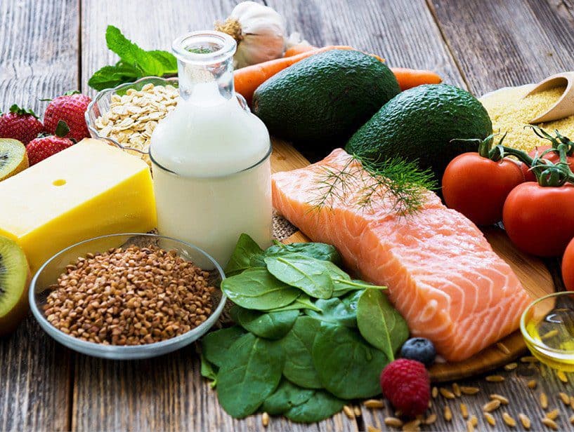 Assorted fresh foods including salmon, avocados, tomatoes, spinach, and dairy on a wooden table.