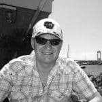 Man wearing sunglasses and cap smiling outdoors near water.