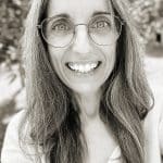 Close-up of woman with glasses smiling outdoors in black and white.