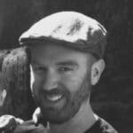 Portrait of Aaron C. wearing a flat cap, smiling at the camera.