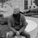 Man sitting on a curved concrete bench outdoors in a park setting.