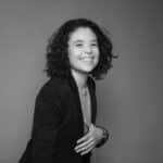 Smiling woman with curly hair in a blazer against a plain background.