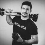Man holding camera and tripod, wearing Virtual Vista T-shirt.