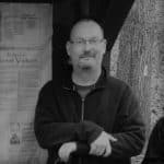 Man with glasses and beard standing outdoors near a tree.