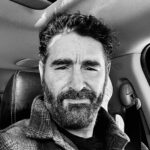 Black and white photo of a bearded man with curly hair inside a car.